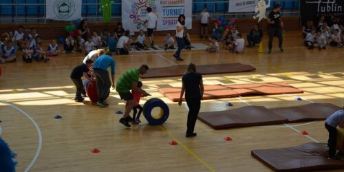 wyścigi rzędów w hali sportowej - dzieci z pomocą nauczycieli pchają beczki rehabilitacyjne